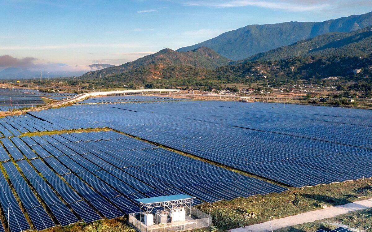 Rows of photovoltaic modules across a utility-scale solar field, with an inverter station in the foreground and hills beyond