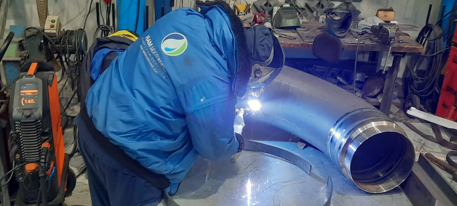 A RAM welder in branded workwear running a TIG weld on a large-bore pipe elbow.