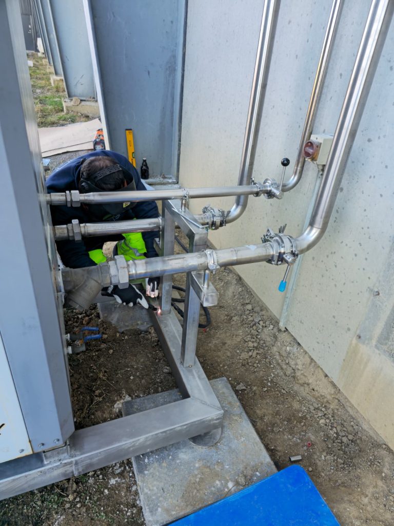 RAM project: Welder working on a stainless steel support frame beside external brewery pipework